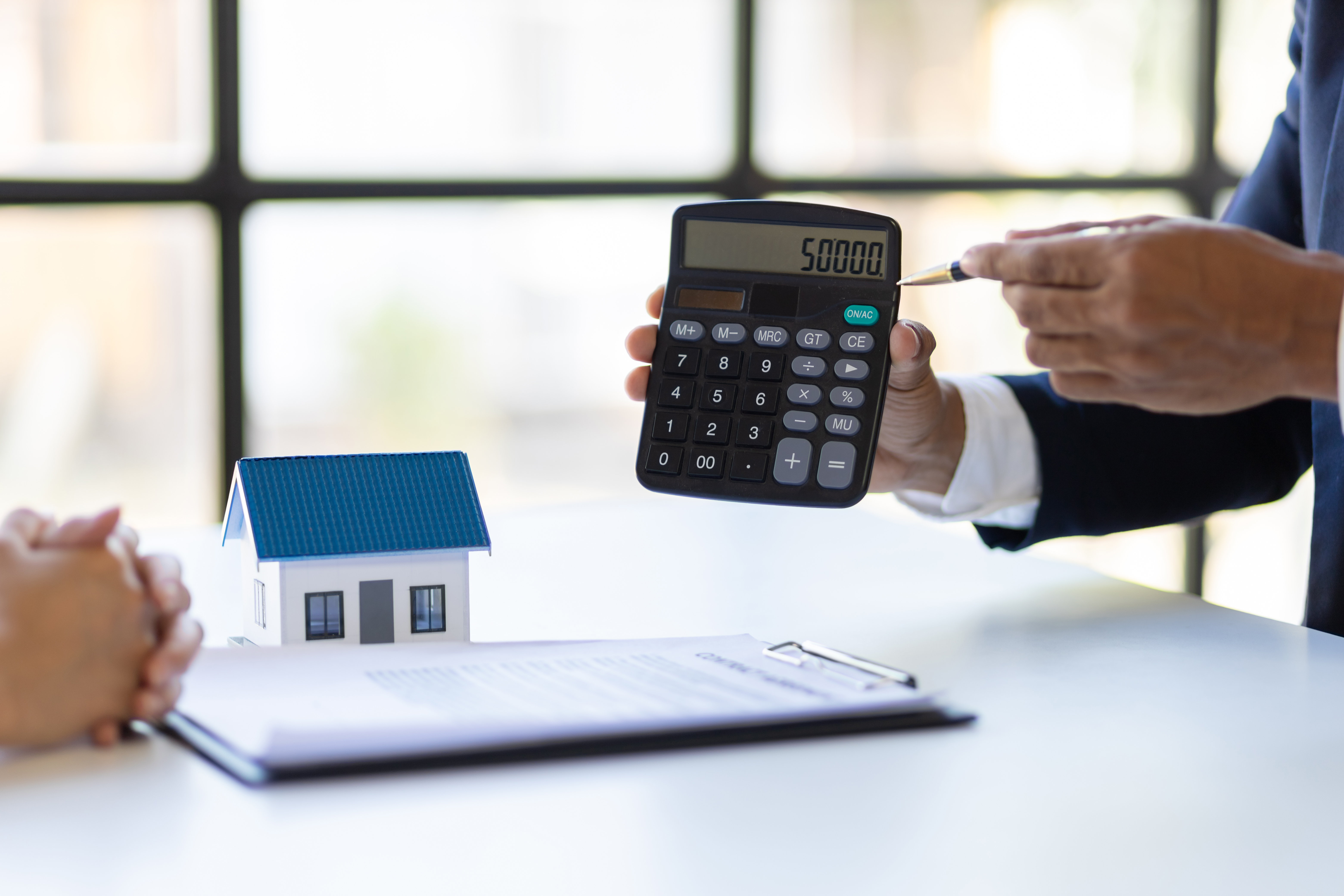 A person holding up a calculator and pointing at it while a document and a figurine of a house sit on the table below
